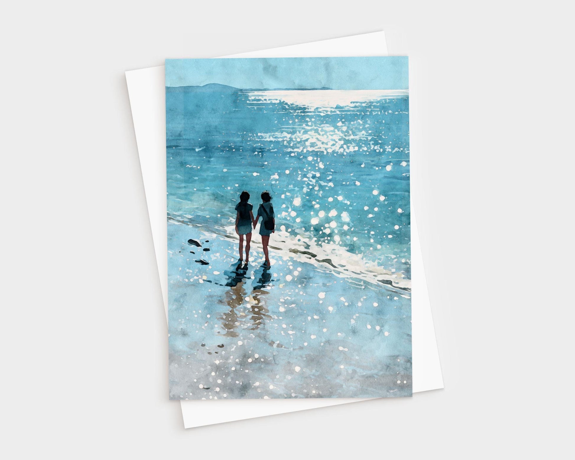 Watercolor-style greeting card featuring two people holding hands while walking along a sparkling shoreline, with their reflections in the wet sand and sunlight glittering on the ocean. A white envelope is partially visible behind the card.