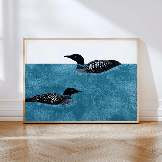 Minimalist illustration of two black and white loons with red eyes swimming in textured blue water, set against a clean white background. The artwork is framed in light wood and displayed leaning against a white wall on a herringbone wood floor, with natural sunlight casting soft shadows across the scene.