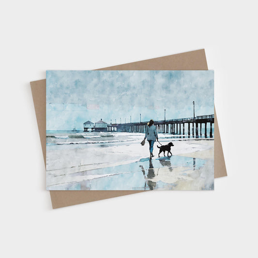 Watercolor-style greeting card featuring a woman walking a black dog along a reflective shoreline, with a long wooden pier stretching into the ocean under a soft, overcast blue sky.