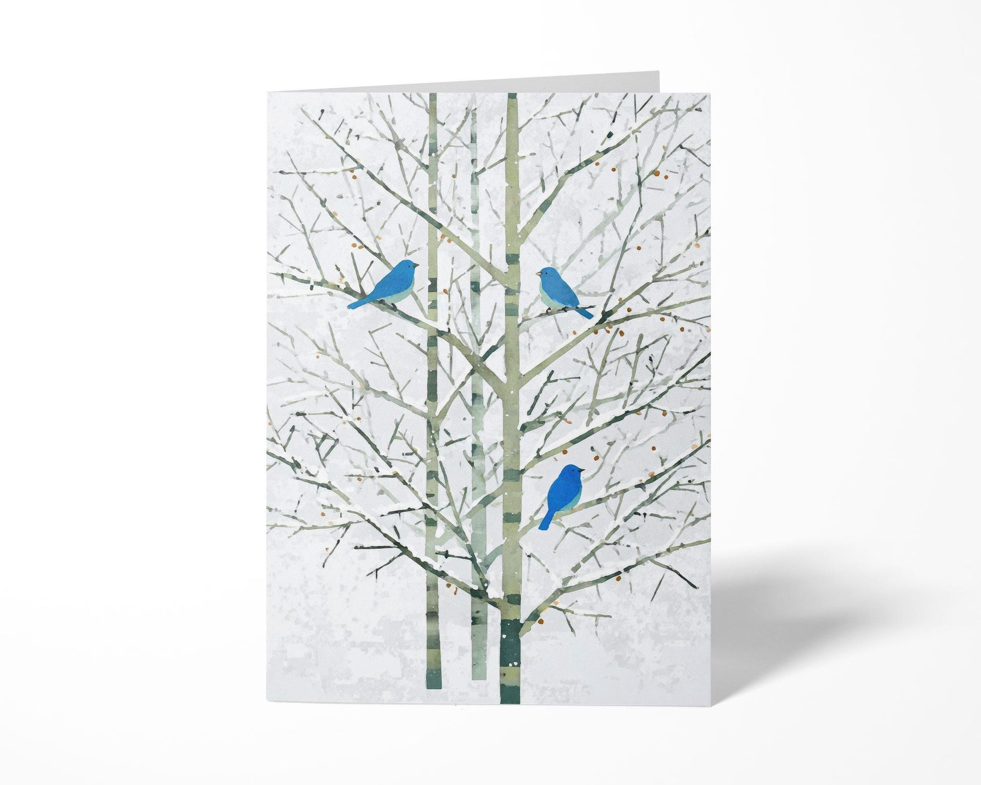 Folded greeting card featuring a minimalist watercolor-style illustration of three bright bluebirds perched on bare winter branches with soft green trunks and a pale white background. The card stands upright on a clean white surface, emphasizing its elegant, modern design.