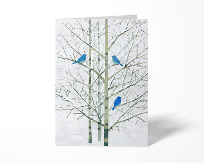 Folded greeting card featuring a minimalist watercolor-style illustration of three bright bluebirds perched on bare winter branches with soft green trunks and a pale white background. The card stands upright on a clean white surface, emphasizing its elegant, modern design.