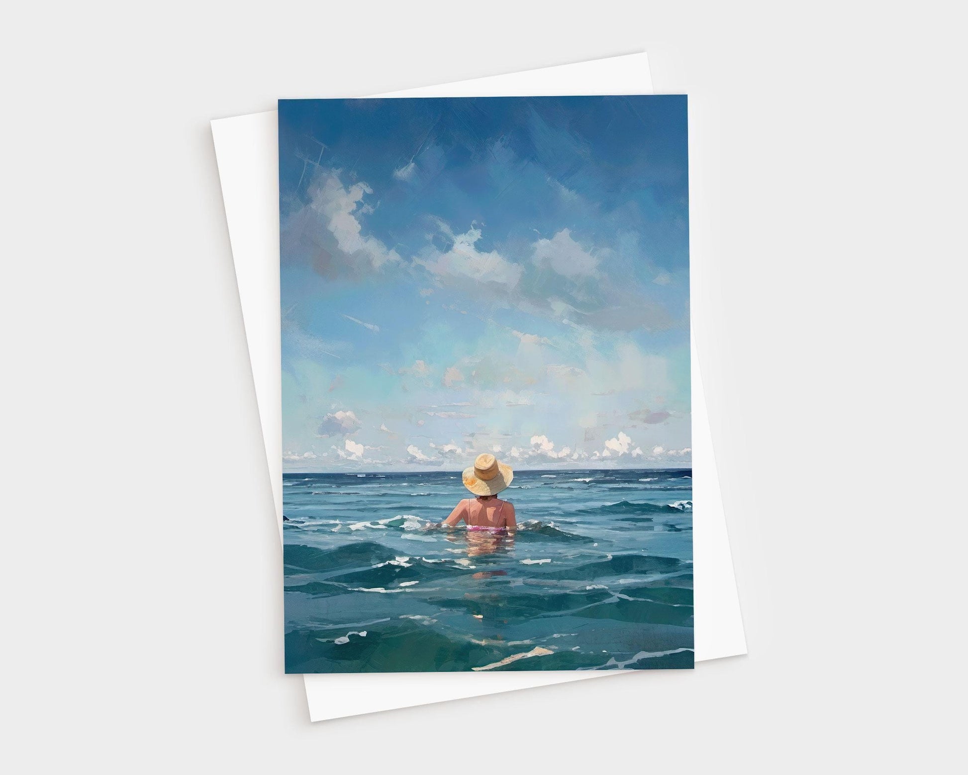 Greeting card showing a woman in a straw hat wading into the ocean under a wide sky with soft clouds, placed over a white envelope on a light gray background.