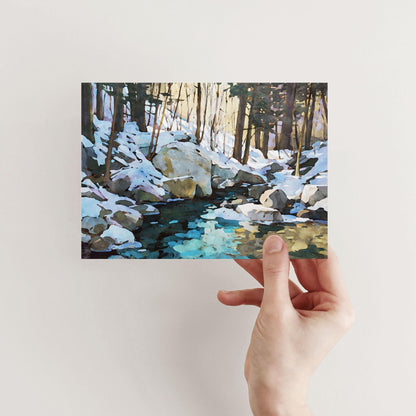 Hand holding a greeting card featuring a watercolor-style snowy forest scene with large rocks and a creek reflecting icy blue and golden tones beneath tall trees.