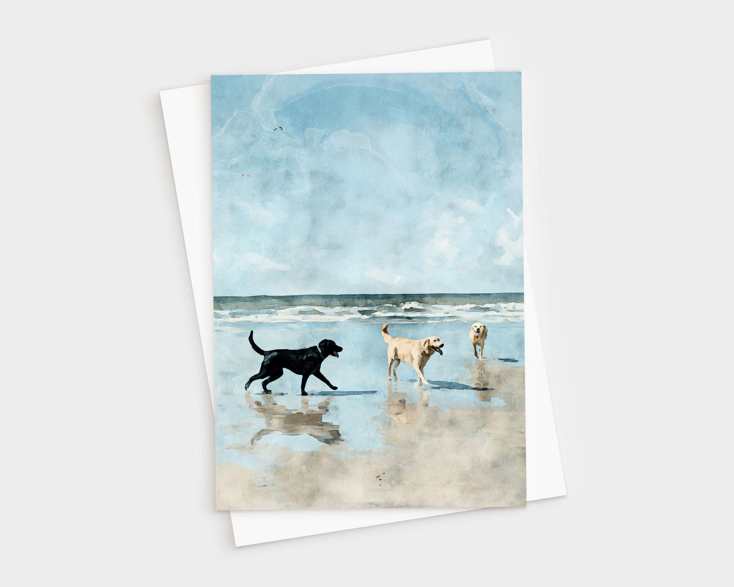 Watercolor-style illustration of three dogs—one black and two golden—playing on a sandy beach with gentle waves and a bright blue sky in the background, printed on a greeting card with a white envelope behind it.