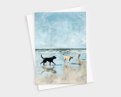 Watercolor-style illustration of three dogs—one black and two golden—playing on a sandy beach with gentle waves and a bright blue sky in the background, printed on a greeting card with a white envelope behind it.