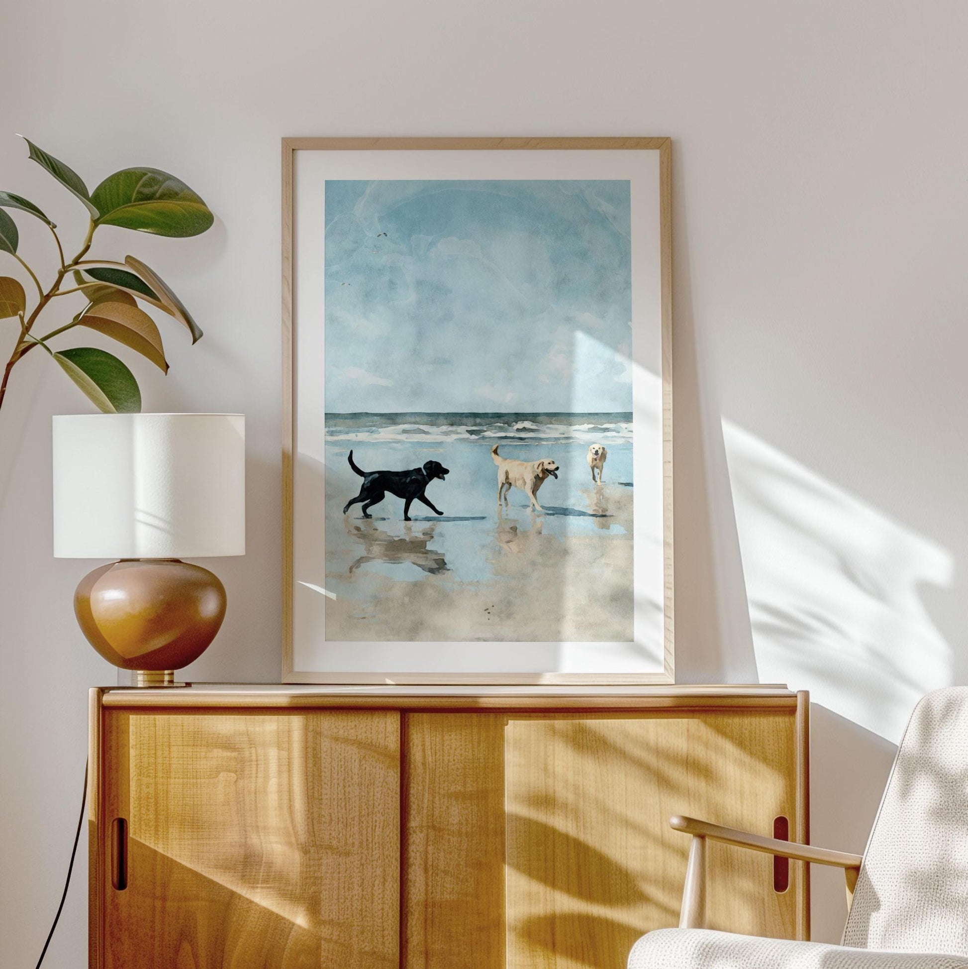Framed watercolor-style print of three Labrador retrievers playing on a beach, hung above a light wood cabinet in a sunlit room. A black lab approaches two yellow labs, one holding a stick, with reflections in wet sand under a blue sky and ocean waves. A ceramic lamp and leafy plant sit on the cabinet, with sunlight casting soft shadows on the wall.
