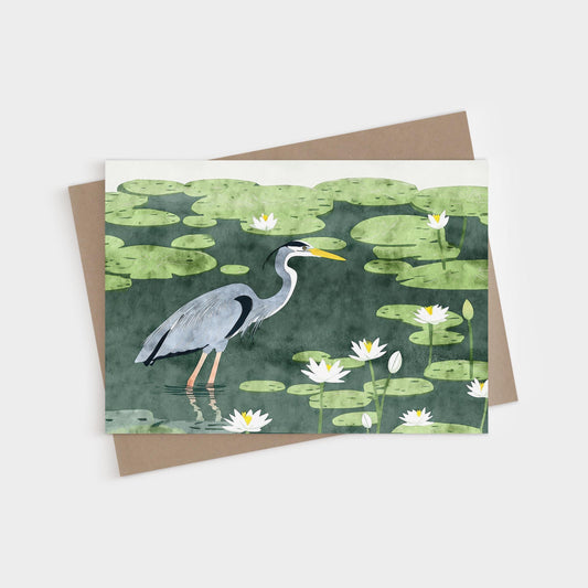 Greeting card featuring an illustration of a great blue heron wading among lily pads and white water lilies in a dark green pond. The heron is shown in profile with detailed feathers and a bright yellow beak. A kraft brown envelope is partially visible beneath the card.