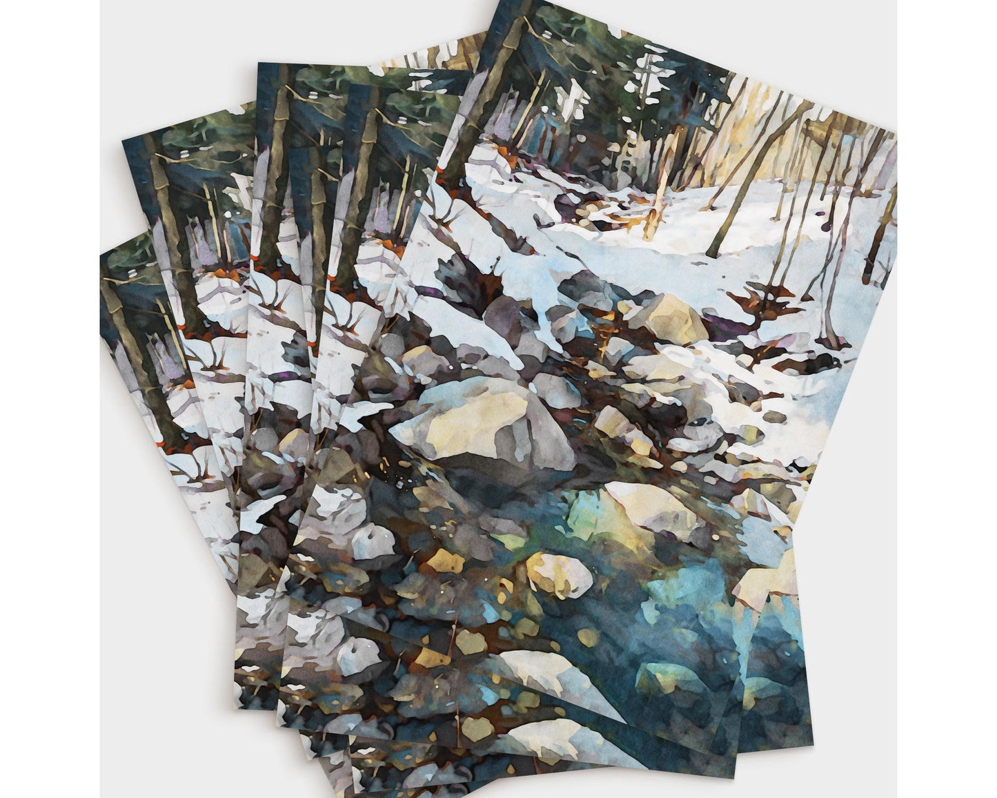 Set of greeting cards featuring a watercolor-style painting of a snowy forest creek with rocks and icy blue-green reflections in the water, fanned out to show multiple copies of the same design.