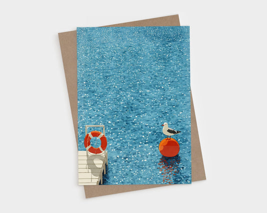 Greeting card featuring a seagull perched on a bright orange buoy in shimmering blue water, with a wooden dock and life preserver visible in the corner. The artwork has a clean, minimalist watercolor style with sparkling highlights across the surface of the water. A kraft envelope is partially visible beneath the card.