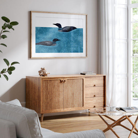 Framed illustration of two loons swimming in textured blue water, hanging above a mid-century modern wooden sideboard with woven cane doors. The bright living room features a white sofa, glass coffee table, and large windows with sheer curtains, creating a light and airy atmosphere with natural greenery visible outside.