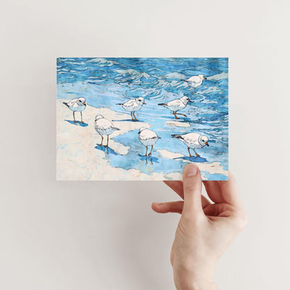 A hand holds a greeting card featuring a watercolor-style illustration of small white shorebirds with orange legs standing and wading along the shoreline, with blue ocean waves in the background.
