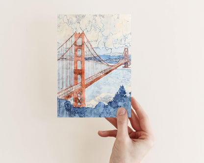 Hand holding a greeting card featuring a watercolor painting of the Golden Gate Bridge in warm rust tones, with soft blue water and fluffy clouds above. The foreground shows loose brushstrokes of navy trees, blending architecture and nature in a light, airy composition.
