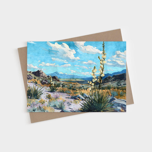 Greeting card featuring a watercolor-style desert landscape with blooming yucca plants, rocky hills, and distant mountains under a vivid blue sky with fluffy white clouds.