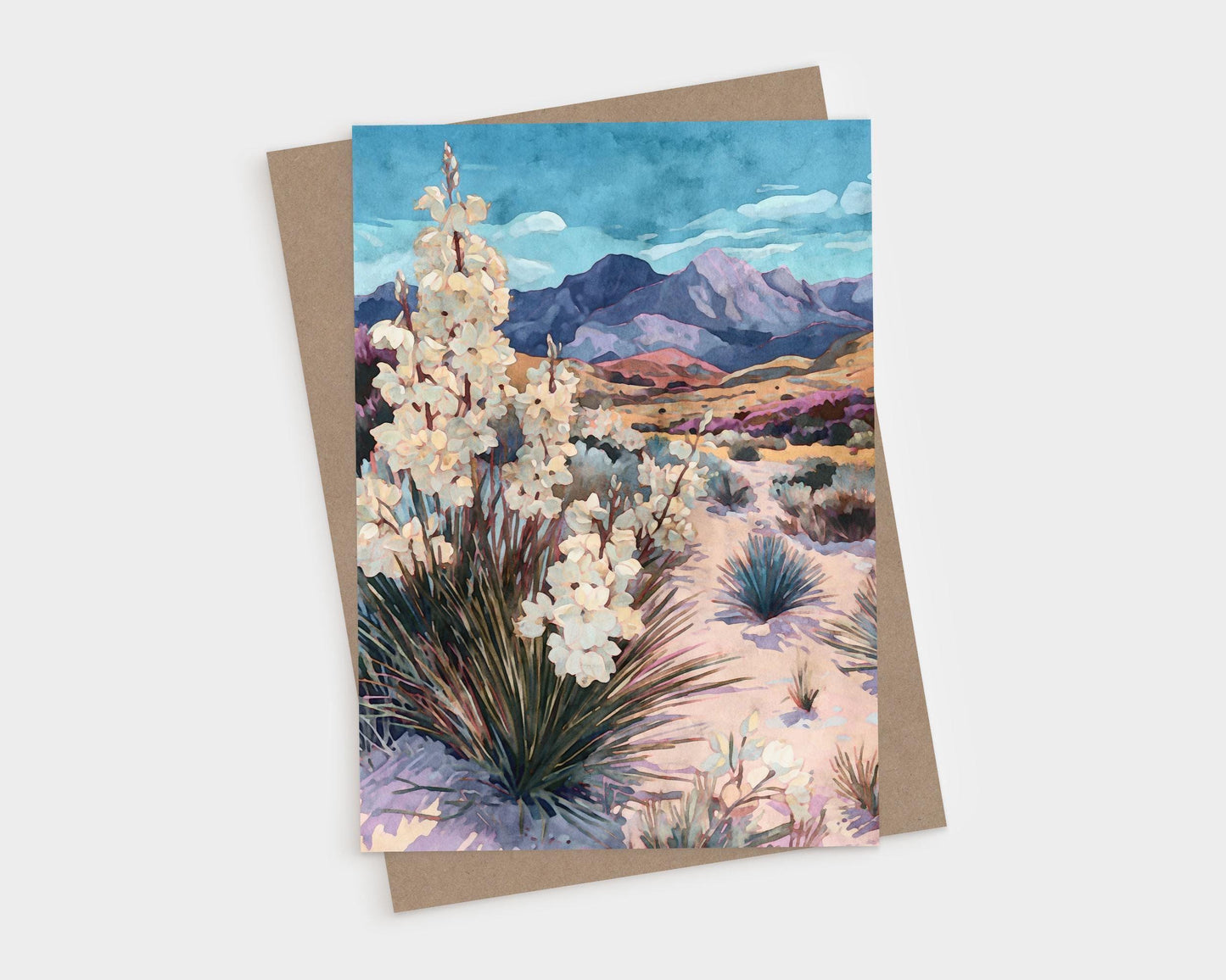 Greeting card featuring a watercolor-style desert landscape with blooming yucca plants in the foreground, sandy terrain with scattered shrubs, and purple mountains under a vivid blue sky, paired with a kraft envelope.