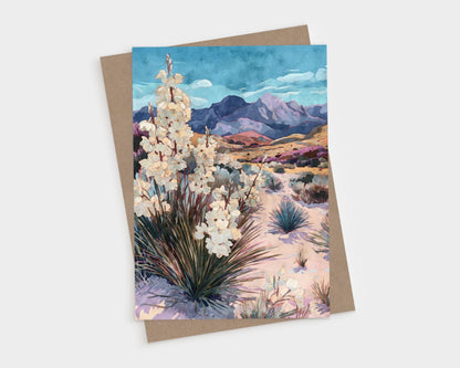 Greeting card featuring a watercolor-style desert landscape with blooming yucca plants in the foreground, sandy terrain with scattered shrubs, and purple mountains under a vivid blue sky, paired with a kraft envelope.