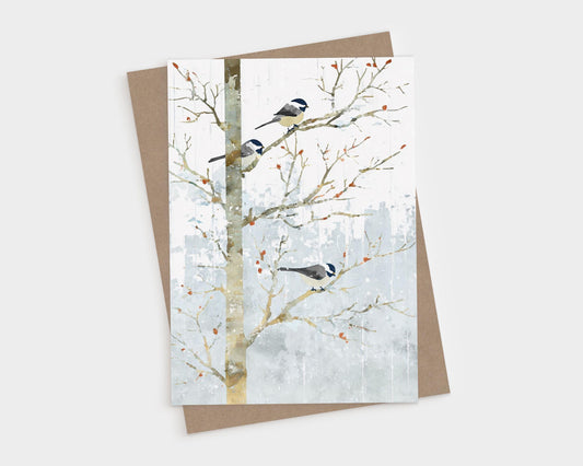 Greeting card featuring a watercolor-style illustration of three chickadees perched on bare winter branches. Soft gray-blue snowy background with scattered orange berries. The card is shown with a Kraft brown envelope behind it on a white surface.