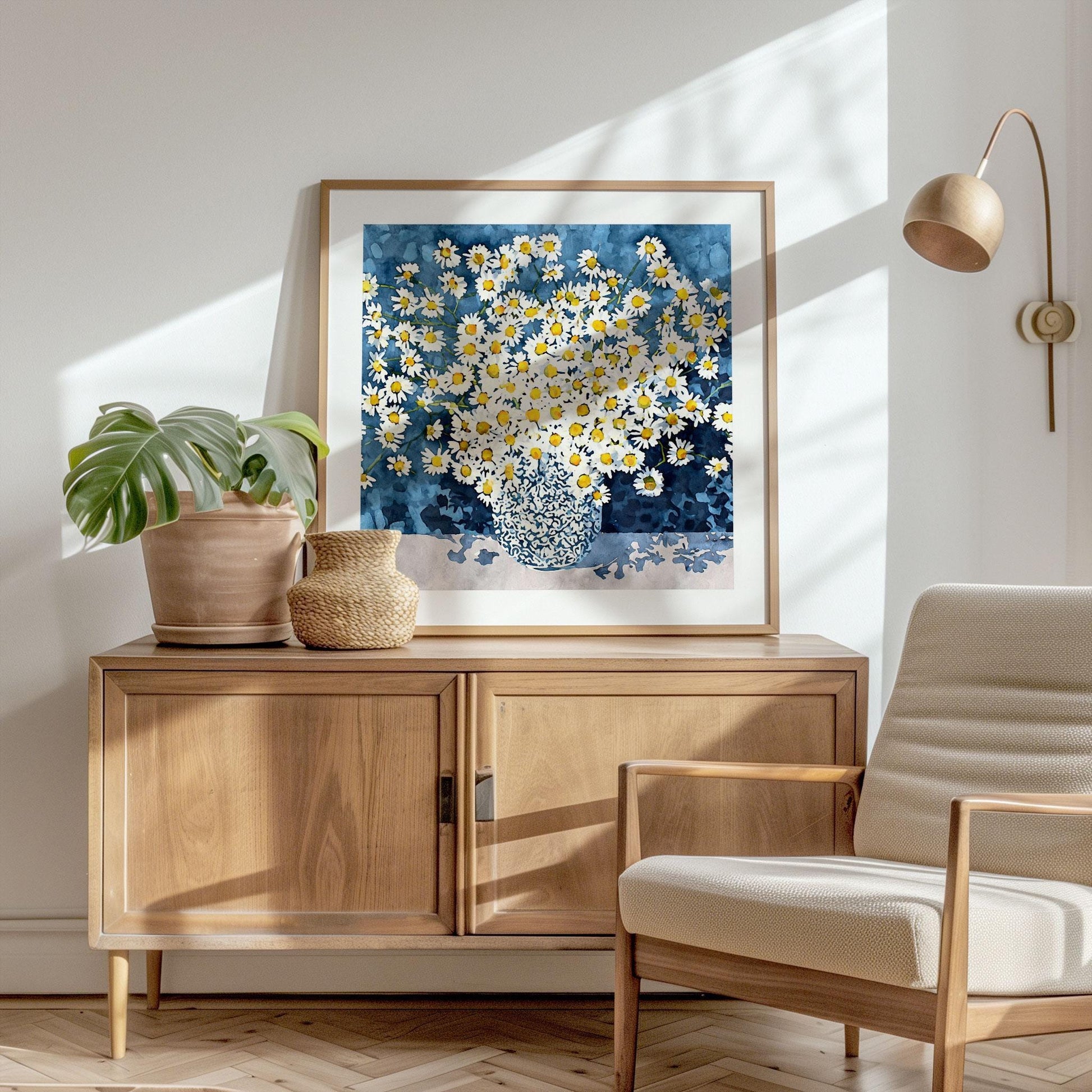 Framed artwork of a bouquet of white daisies with yellow centers in a patterned vase, set against a rich blue background. The print rests on a wooden sideboard, styled with a potted monstera plant and woven basket, in a bright modern room with natural light, a neutral armchair, and a wall-mounted lamp.