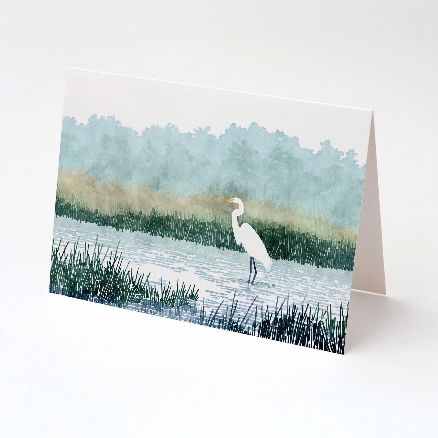 Folded greeting card featuring a watercolor-style illustration of a white heron standing in a marsh with tall grasses, calm water, and misty blue-green trees in the background, displayed against a plain white surface.