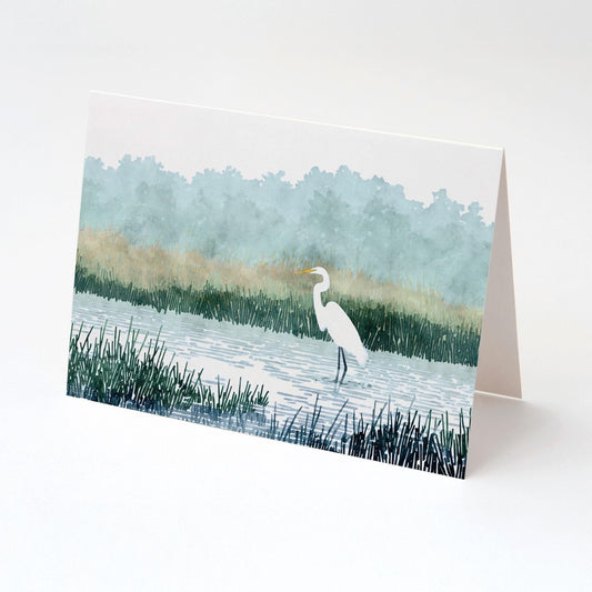 Folded greeting card featuring a watercolor-style illustration of a white heron standing in a marsh with tall grasses, calm water, and misty blue-green trees in the background, displayed against a plain white surface.