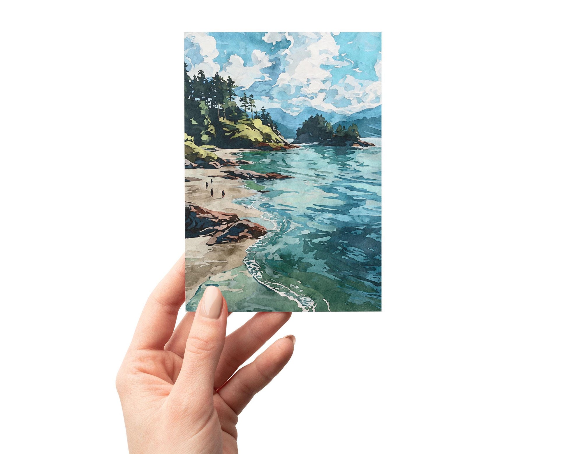 Hand holding a greeting card featuring a watercolor-style coastal landscape with turquoise waves, sandy beach, rocky shoreline, and pine-covered cliffs beneath a bright blue sky with fluffy white clouds.