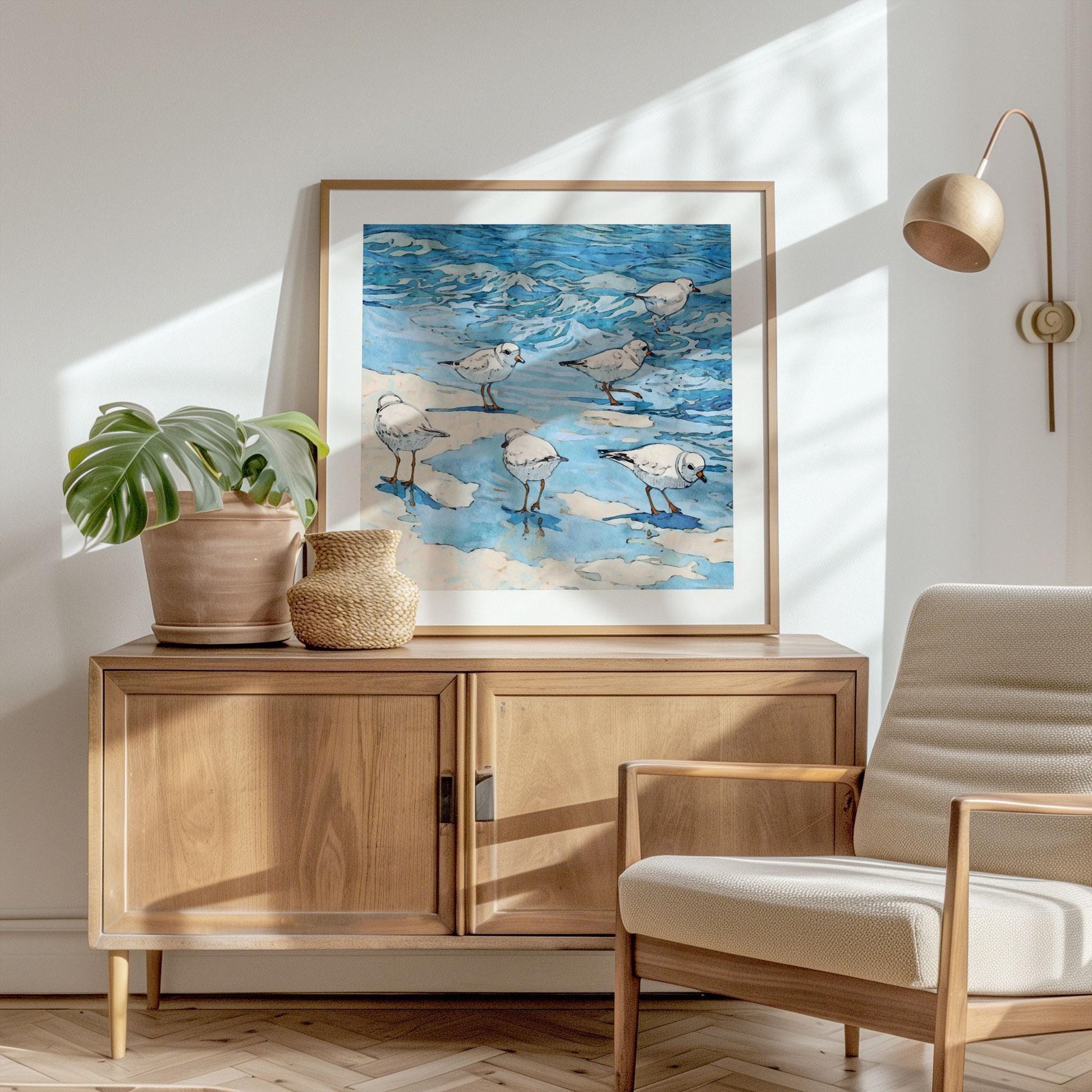 Framed coastal watercolor print depicting a group of small white shorebirds with orange beaks and legs, standing and wading in shallow blue water with soft sandy patches. The waves are rendered in fluid shades of blue with black ink outlines for a lively, illustrative style. Displayed atop a light wood cabinet with potted plants in a sunlit, minimalist room featuring a neutral armchair and wall-mounted lamp.
