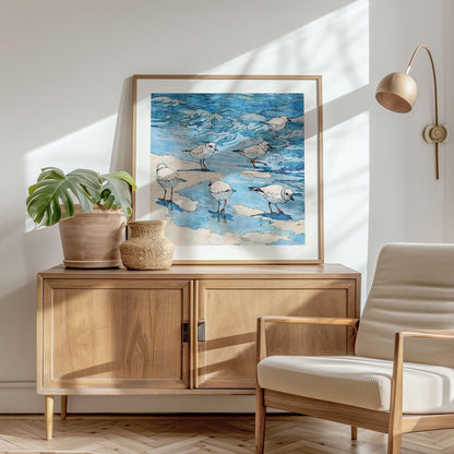 Framed coastal watercolor print depicting a group of small white shorebirds with orange beaks and legs, standing and wading in shallow blue water with soft sandy patches. The waves are rendered in fluid shades of blue with black ink outlines for a lively, illustrative style. Displayed atop a light wood cabinet with potted plants in a sunlit, minimalist room featuring a neutral armchair and wall-mounted lamp.