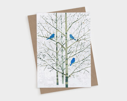 Greeting card featuring a minimalist watercolor-style illustration of three bright bluebirds perched on bare winter branches with pale green trunks and a soft white background. The card is shown with a brown kraft envelope, creating a natural, elegant presentation.
