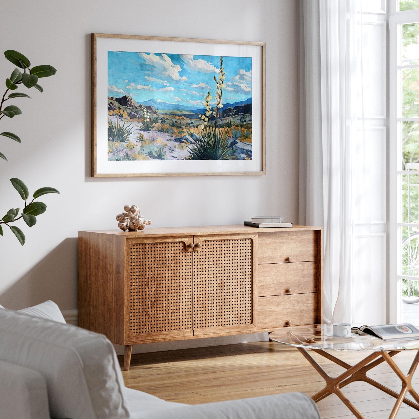 Framed desert landscape painting of yucca plants, rocky terrain, and mountains under a bright blue sky with scattered clouds, displayed above a wooden console in a modern living room with natural light from large windows.