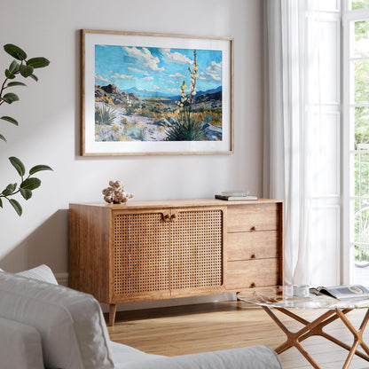 Framed desert landscape painting of yucca plants, rocky terrain, and mountains under a bright blue sky with scattered clouds, displayed above a wooden console in a modern living room with natural light from large windows.