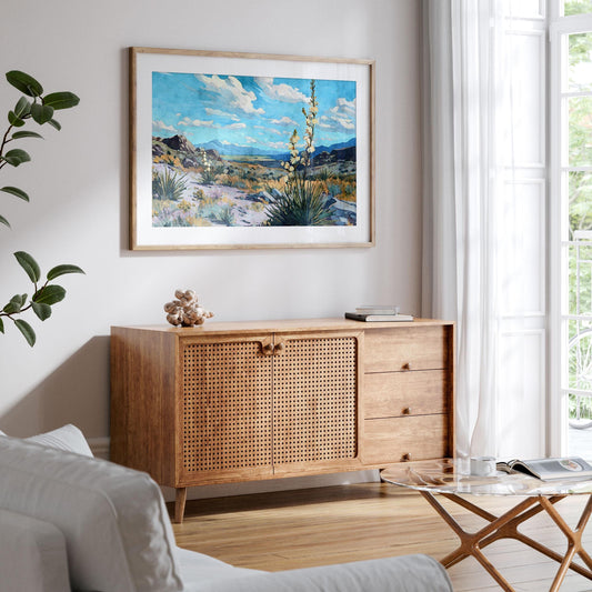 Framed desert landscape painting of yucca plants, rocky terrain, and mountains under a bright blue sky with scattered clouds, displayed above a wooden console in a modern living room with natural light from large windows.