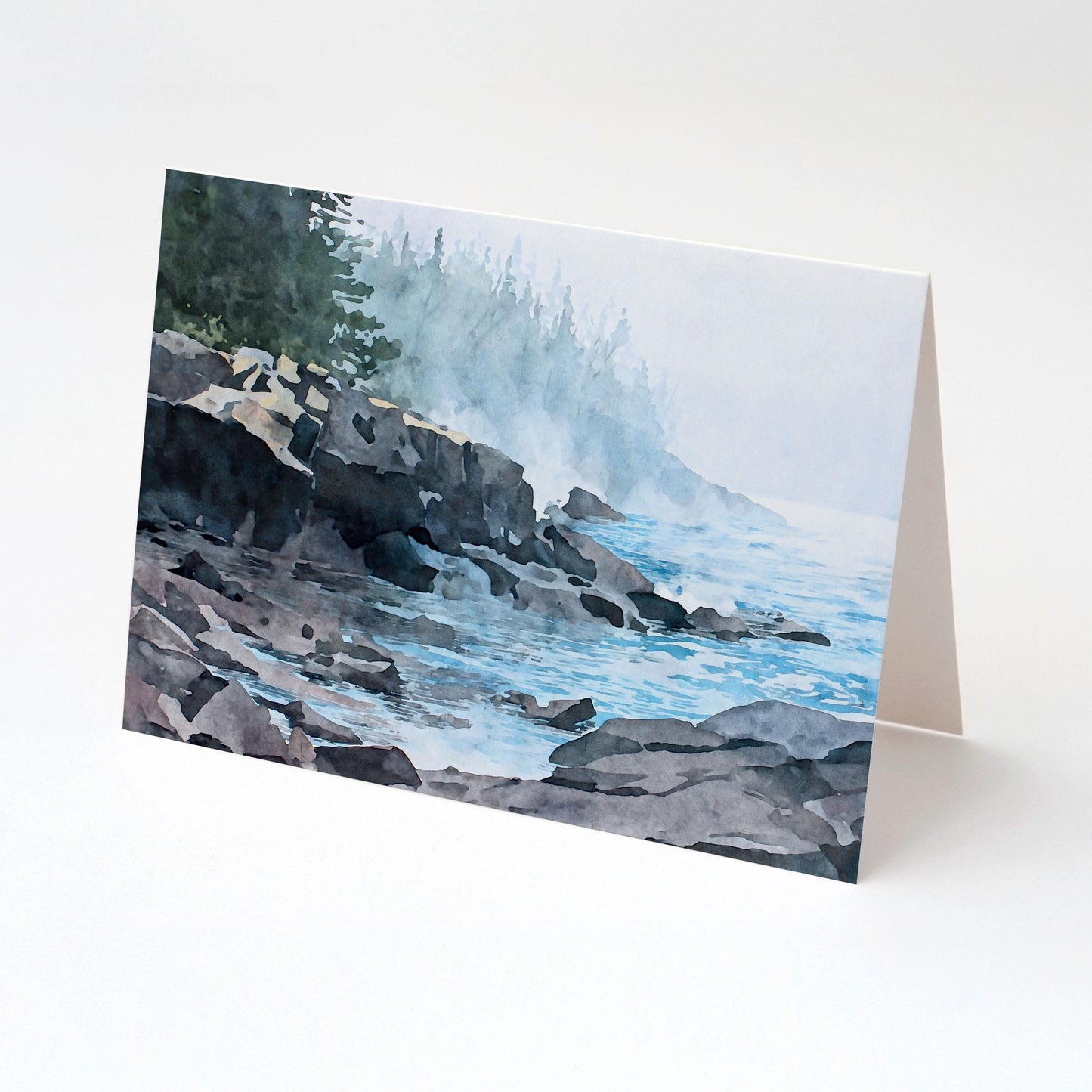 Folded horizontal greeting card standing upright on a white background, featuring a watercolor-style illustration of a misty Lake Superior shoreline. Dark, layered rock formations extend into calm blue water with gentle ripples. Soft fog drifts along the coast, partially obscuring a distant evergreen forest, creating a tranquil, atmospheric lakeside scene in muted blues, grays, and greens.