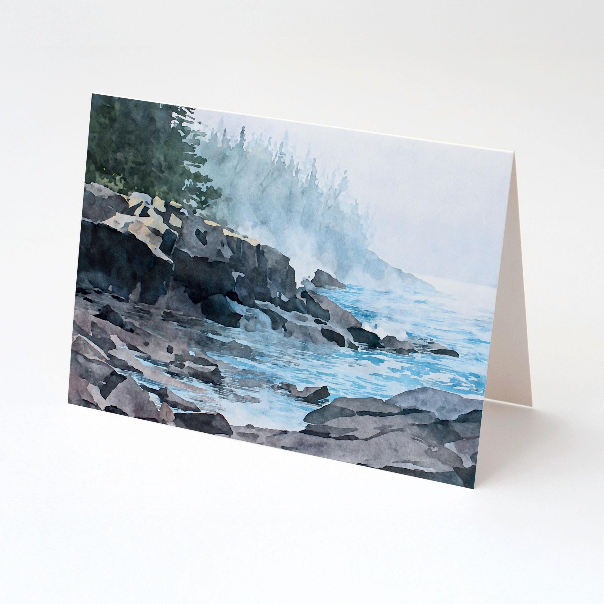 Folded horizontal greeting card standing upright on a white background, featuring a watercolor-style illustration of a misty Lake Superior shoreline. Dark, layered rock formations extend into calm blue water with gentle ripples. Soft fog drifts along the coast, partially obscuring a distant evergreen forest, creating a tranquil, atmospheric lakeside scene in muted blues, grays, and greens.