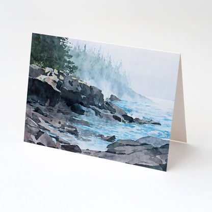 Folded horizontal greeting card standing upright on a white background, featuring a watercolor-style illustration of a misty Lake Superior shoreline. Dark, layered rock formations extend into calm blue water with gentle ripples. Soft fog drifts along the coast, partially obscuring a distant evergreen forest, creating a tranquil, atmospheric lakeside scene in muted blues, grays, and greens.
