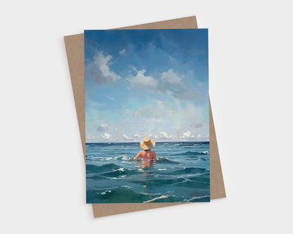 Greeting card showing a woman in a straw hat wading into the ocean under a wide sky with soft clouds, placed over a kraft envelope on a light gray background.