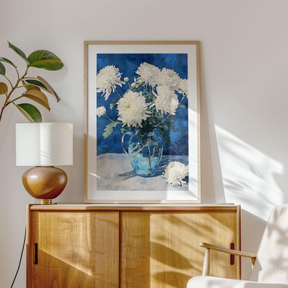 Framed art print of white chrysanthemums in a glass pitcher with a deep blue background, styled on a light wood cabinet. A modern ceramic lamp and leafy plant add warmth to the minimalist room, with sunlight casting soft shadows on the wall.