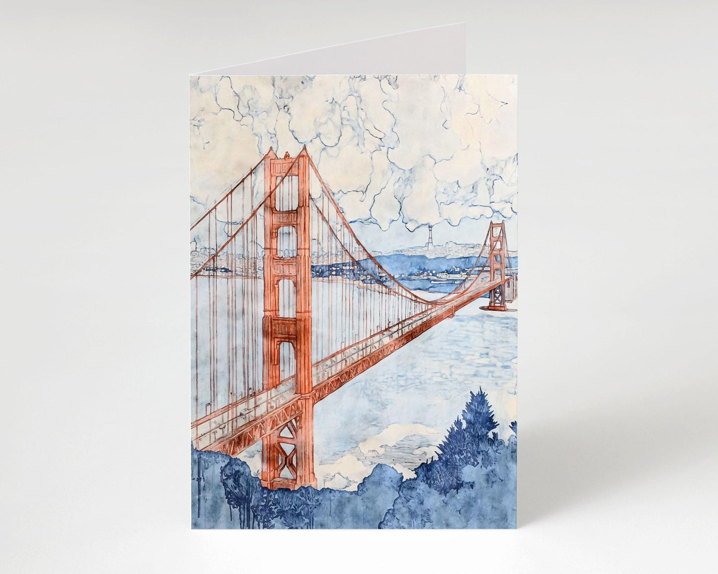 Greeting card featuring a watercolor painting of the Golden Gate Bridge in shades of rust red and cool blue. The bridge stretches across a tranquil bay beneath a cloudy sky, with soft blue foliage in the foreground and distant hills in the background. The card stands open against a clean, light backdrop.