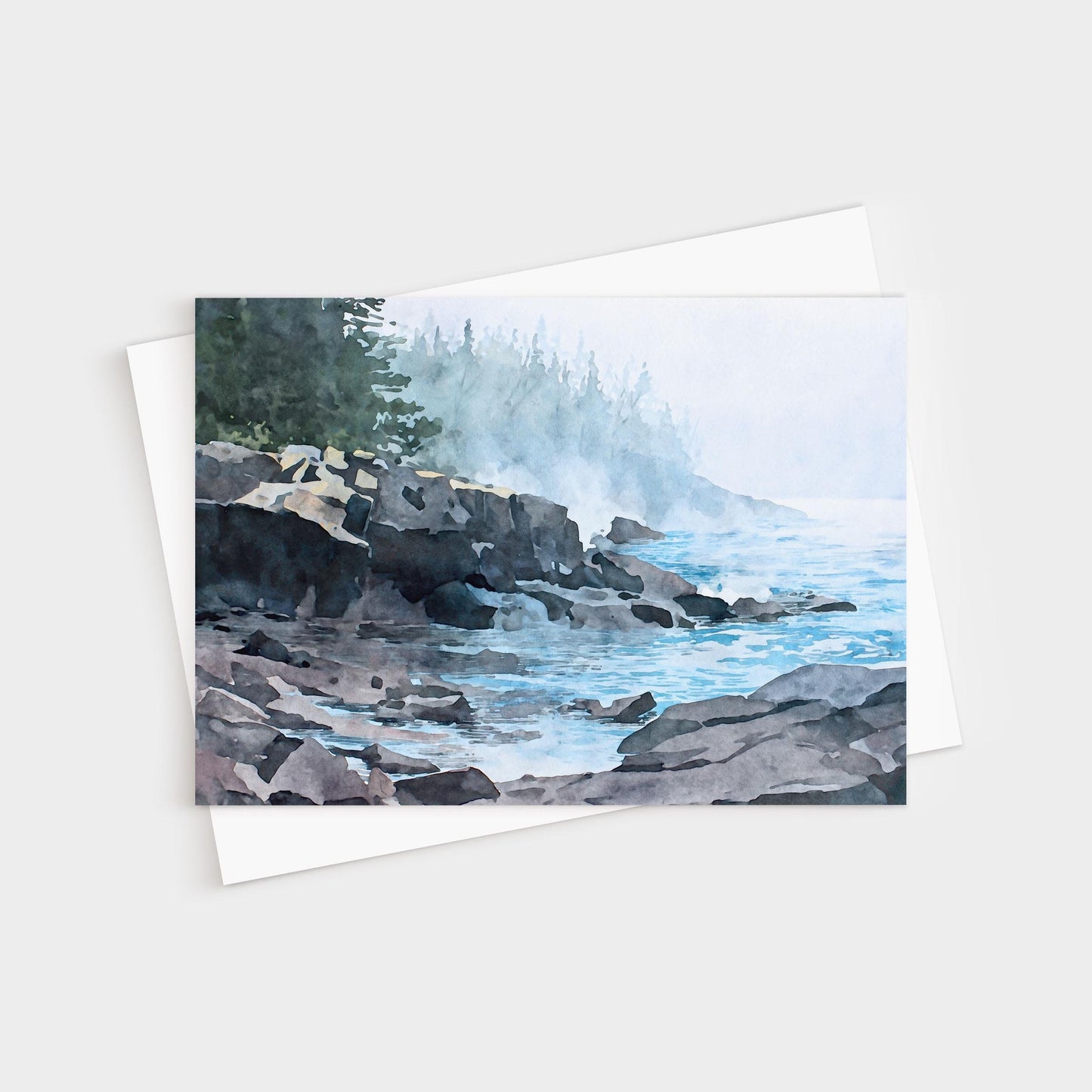 Horizontal greeting card featuring a watercolor-style illustration of a misty Lake Superior shoreline. Dark, layered rocky cliffs extend into calm blue water, with gentle waves lapping against the stones. A soft fog drifts across the scene, partially obscuring a distant forest of evergreen trees. Muted blues, grays, and greens create a quiet, atmospheric coastal mood.
