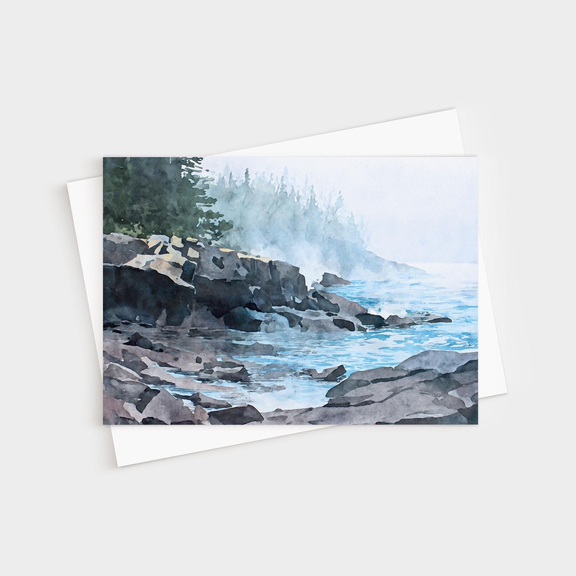 Horizontal greeting card featuring a watercolor-style illustration of a misty Lake Superior shoreline. Dark, layered rocky cliffs extend into calm blue water, with gentle waves lapping against the stones. A soft fog drifts across the scene, partially obscuring a distant forest of evergreen trees. Muted blues, grays, and greens create a quiet, atmospheric coastal mood.