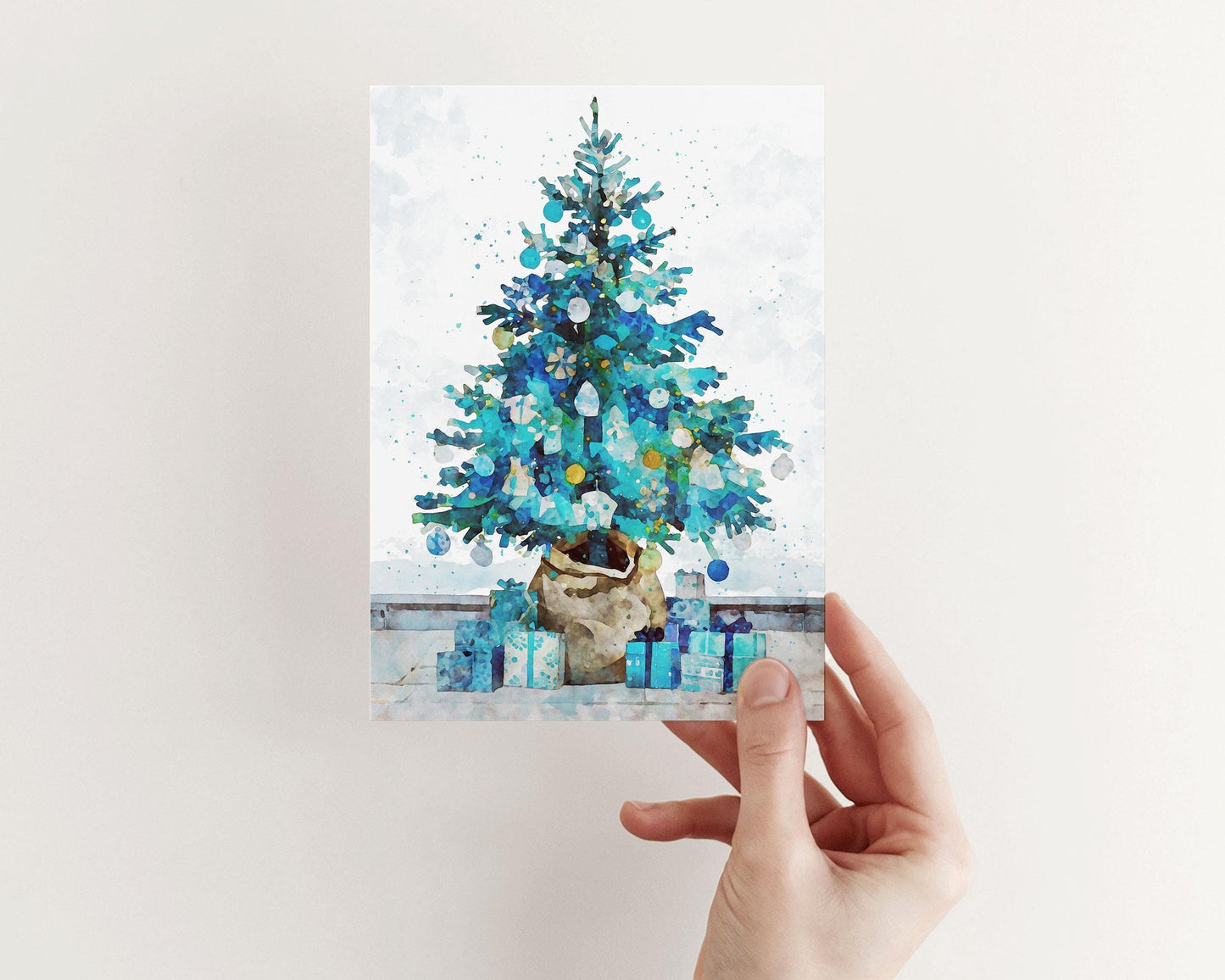 Hand holding a watercolor-style Christmas card featuring a blue and teal holiday tree in a burlap-wrapped pot, surrounded by matching blue presents on a pale background.