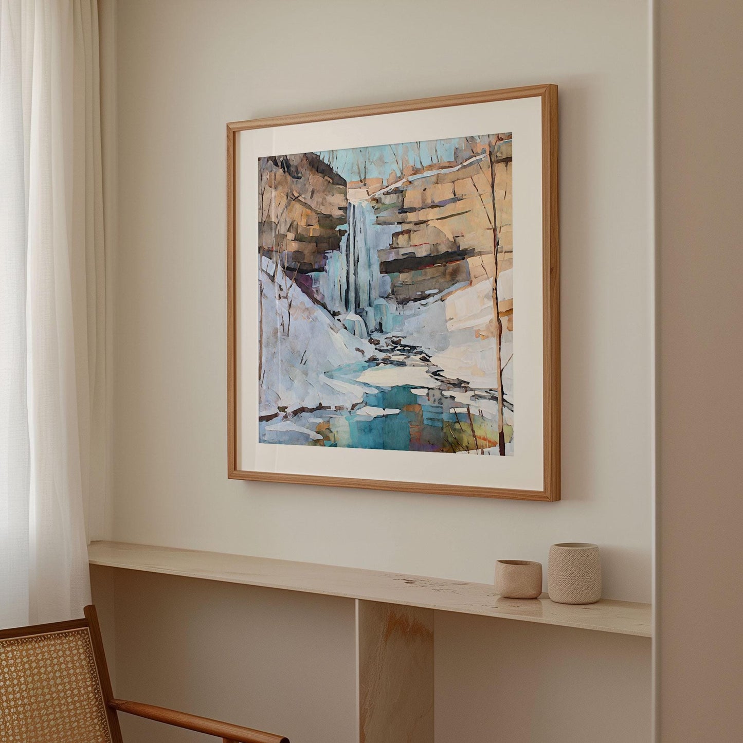 Framed watercolor painting of a snowy waterfall and turquoise pool, hanging on a neutral wall above a light wood shelf with minimalist ceramic decor, in a softly lit modern room.