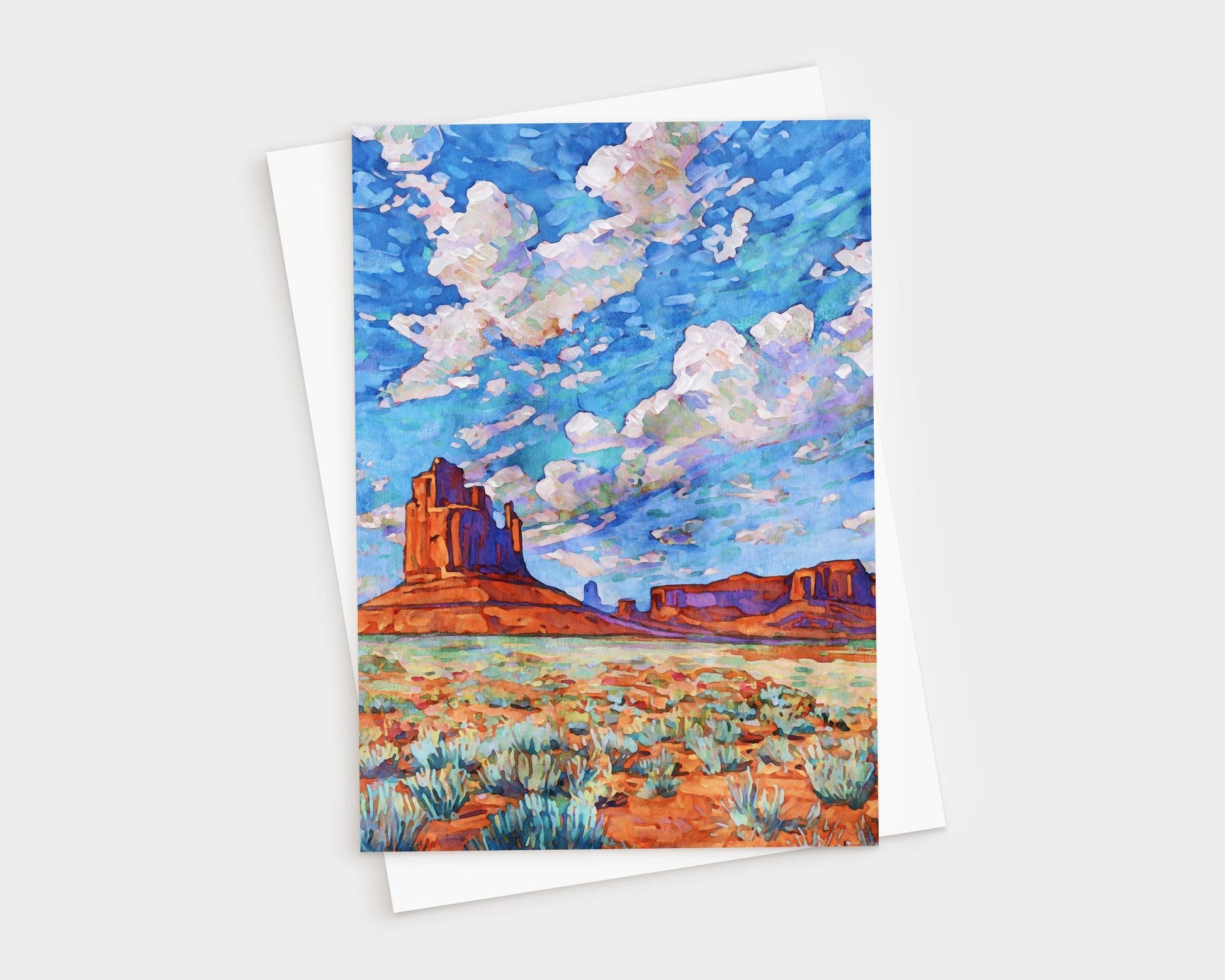 Vibrant greeting card featuring a painting of Monument Valley. The scene shows towering red rock buttes rising from a desert landscape dotted with green sagebrush. Above, a dramatic sky of vivid blues and purples is filled with fluffy white clouds, painted in bold, impressionistic strokes. The artwork captures the vast, colorful beauty of the American Southwest.