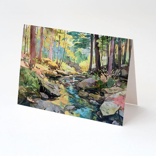 Folded greeting card featuring a watercolor woodland scene with tall trees, scattered rocks, and a stream reflecting sunlight. The card is displayed upright against a plain white background, highlighting the vivid greens, blues, and yellows of the artwork.