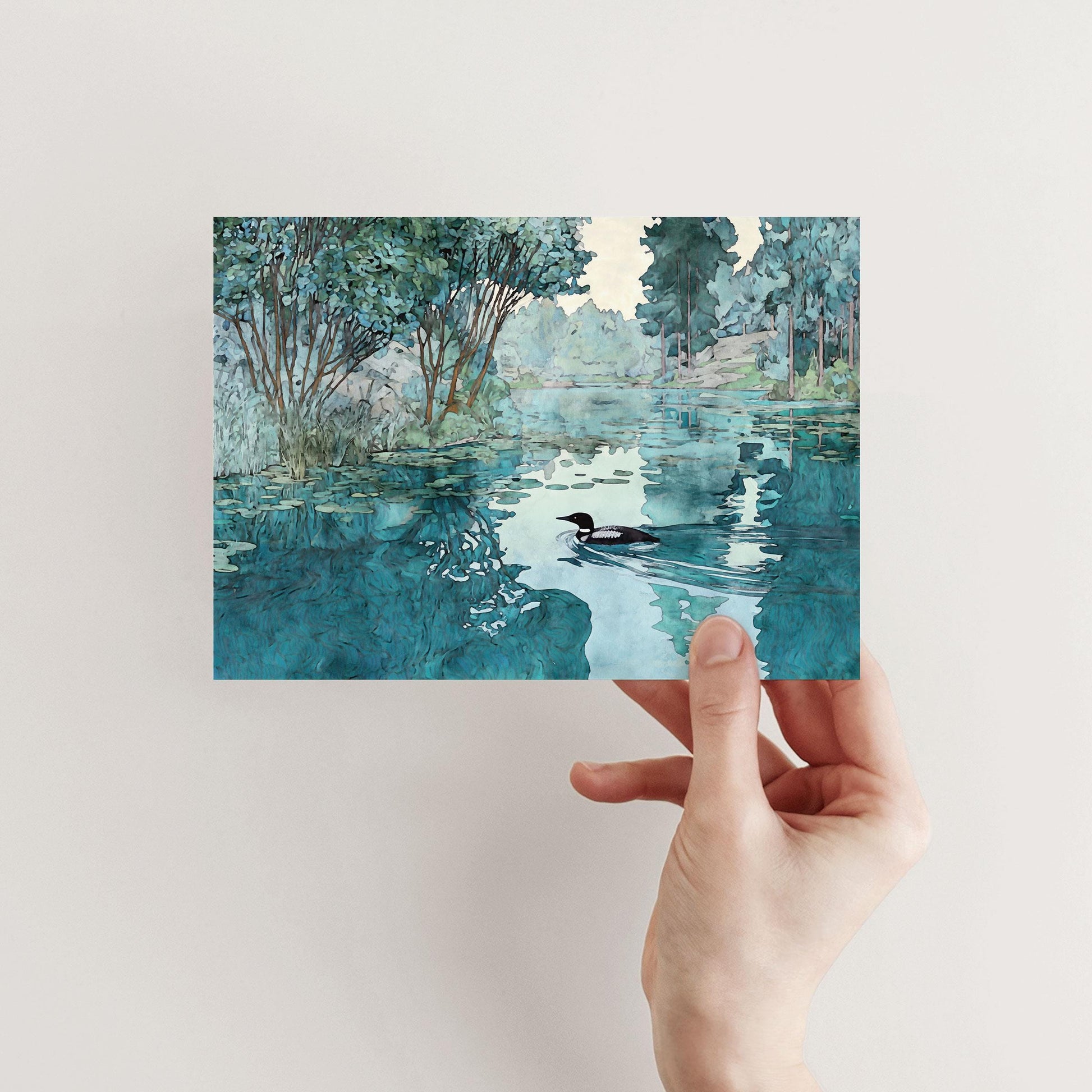 A hand holds a greeting card featuring a watercolor-style illustration of a loon swimming on a calm lake. The scene includes reflections of green trees, shrubs, and a soft blue sky, creating a peaceful, nature-inspired design.