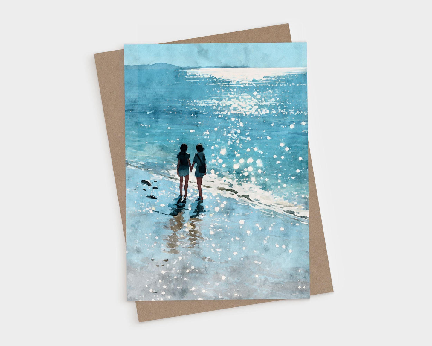 Watercolor-style greeting card featuring two people holding hands while walking along a sparkling shoreline, with their reflections in the wet sand and sunlight glittering on the ocean. A kraft envelope is partially visible behind the card.