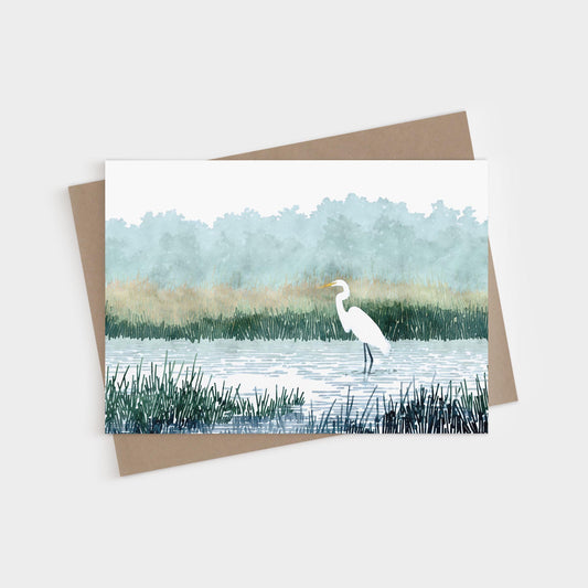 Watercolor-style illustration of a white heron standing in a marsh, surrounded by tall grasses and calm water, with soft green trees and golden reeds in the misty background. The card is displayed with a brown kraft envelope.