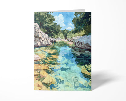Folded greeting card featuring a watercolor-style painting of a turquoise river with golden rocks beneath the clear water, surrounded by rocky white banks and lush green trees under a blue sky with clouds. Displayed upright on a plain white background.