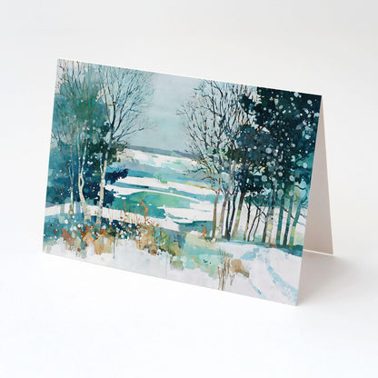 Folded greeting card standing upright, featuring a watercolor winter landscape of snow-covered fields, bare trees, and evergreens in teal, blue, and white tones with splattered paint details suggesting falling snow.