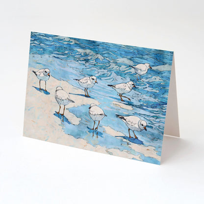 Folded greeting card featuring a watercolor-style illustration of small white shorebirds with orange legs on sand and shallow blue water, displayed upright against a clean white background.