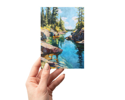 Hand holding a greeting card featuring a watercolor painting of a canoe floating in a narrow turquoise lake surrounded by rocky cliffs and tall pine trees under a bright blue sky with clouds.