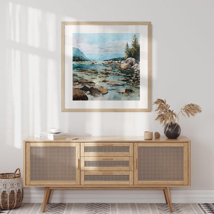 Framed watercolor painting of a rocky mountain river hangs above a mid-century style cane-front sideboard in a bright, minimalist room. The artwork depicts turquoise water flowing through stones toward distant blue mountains and pine trees under a pastel sky. Decor includes a small black vase with dried grass, neutral ceramics, and a woven basket, all softly lit by natural sunlight.